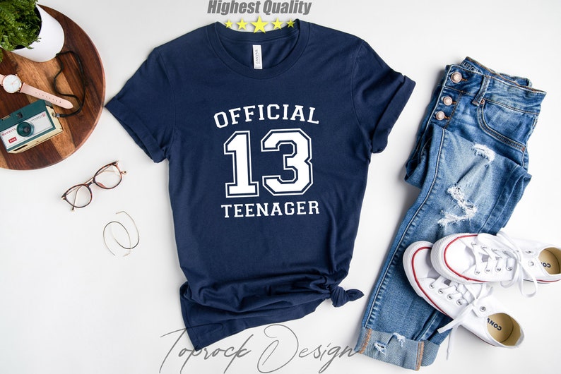 May include: A navy blue t-shirt with white text that reads "OFFICIAL 13 TEENAGER". The shirt is laid flat on a white surface with a pair of jeans and white trainers.