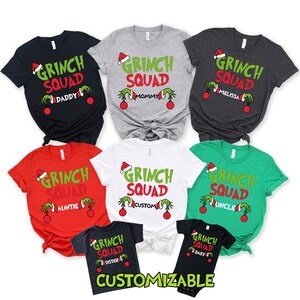 May include: A collection of Christmas-themed t-shirts in various colors, including black, gray, red, white, and green. Each shirt features the text "GRINCH SQUAD" with different family member names or customizable options, along with Grinch-themed graphics.