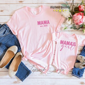May include: Pink t-shirts and a baby onesie with the text "MAMA est. 2025" and "MAMA'S girl" in pink. The shirts are paired with blue jeans, ballet flats, and baby shoes. The image is on a light wood background.