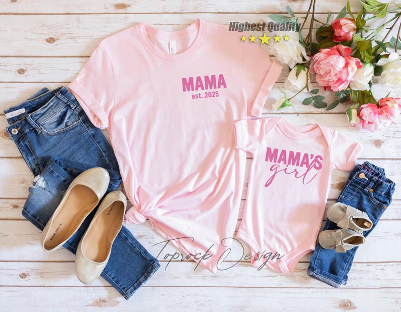May include: Pink t-shirts and a baby onesie with the text "MAMA est. 2025" and "MAMA'S girl" in pink. The shirts are paired with blue jeans, ballet flats, and baby shoes. The image is on a light wood background.