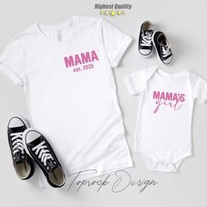 May include: White t-shirts and a baby onesie with pink text that reads "MAMA est. 2025" and "MAMA'S girl". Black and white sneakers are placed near the shirts. The image is a flat lay on a light gray surface.