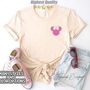 Puede incluir: Una camiseta beige claro con una silueta rosa de Minnie Mouse en la parte delantera. La camiseta est&aacute; sobre una superficie blanca con un par de zapatos Converse rosas y unos pantalones cortos de mezclilla azul con gafas de sol marrones.