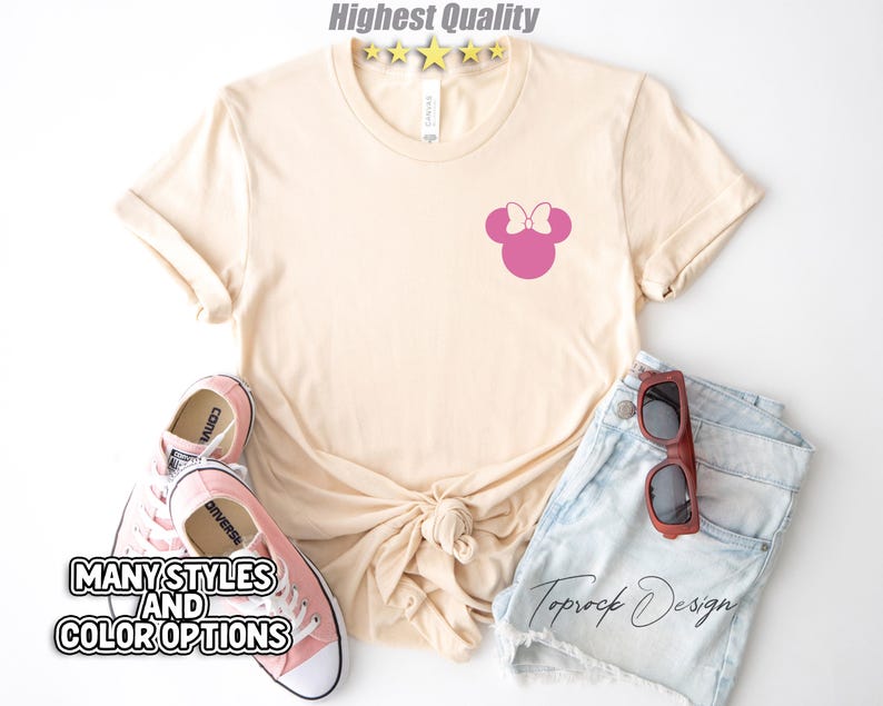 Puede incluir: Una camiseta beige claro con una silueta rosa de Minnie Mouse en la parte delantera. La camiseta est&aacute; sobre una superficie blanca con un par de zapatos Converse rosas y unos pantalones cortos de mezclilla azul con gafas de sol marrones.
