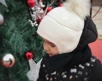 Puede incluir: Un niño lleva un gorro blanco de punto con un pompón esponjoso y una chaqueta negra con un patrón blanco. El niño está mirando un árbol de Navidad decorado con adornos rojos.