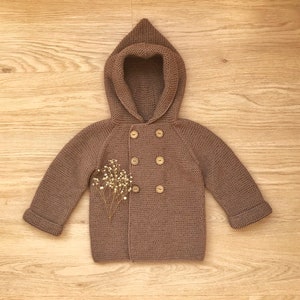 May include: A brown knitted hooded cardigan with a double breasted button closure. The cardigan has a small bouquet of dried flowers on the front.