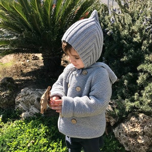 Puede incluir: Una chaqueta con capucha y gorro a juego de punto gris claro con una parte superior puntiaguda. La chaqueta tiene botones de madera y una capucha. El niño lleva pantalones azul oscuro y zapatos grises. El niño sostiene una hoja marrón.