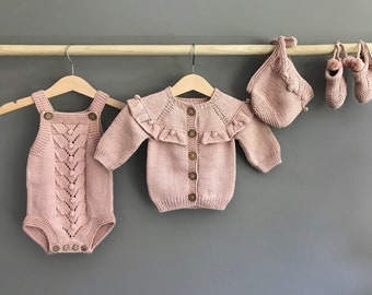 May include: A set of baby clothes in a pale pink color, including a knitted cardigan with a ruffle collar, a knitted romper with a button closure, a knitted hat, and a pair of knitted booties.
