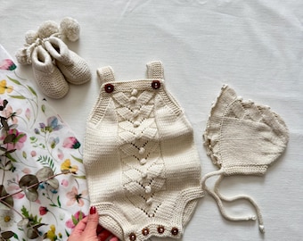 May include: A cream-colored knitted baby romper with a matching bonnet and booties. The romper features a decorative pattern and brown buttons. The set is displayed on a floral patterned cloth.