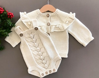 May include: A cream-colored knitted baby outfit set with a cardigan and a romper. The cardigan has a ruffled collar and the romper has a decorative pattern with small buttons.