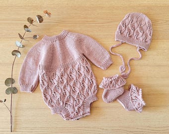 May include: A knitted baby set in a soft rose color, including a romper, hat, and booties. The romper features a detailed leaf pattern on the lower half. The hat has ear flaps and ties, and the booties have a ruffled edge.