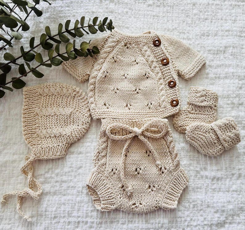 May include: A knitted baby outfit set in a cream color, including a cardigan, pants, hat, and booties. The cardigan has a button closure and a cable knit pattern. The pants have a bow tie detail and a lace pattern. The hat is a simple beanie style. The booties are a simple sock style.