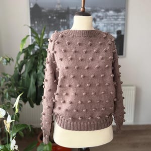 Puede incluir: Un suéter de punto color malva con cuello redondo y detalles de bolitas. El suéter se exhibe en un maniquí, mostrando su textura y diseño. Las bolitas están espaciadas uniformemente en la parte delantera y las mangas.