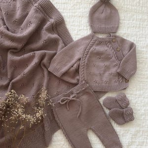 Könnte beinhalten: Ein gestricktes Baby-Set in gedämpftem Mauve, bestehend aus einer Decke, einer Mütze mit Bommel, einem Cardigan mit Holzknöpfen, einer Hose mit Kordelzug und Babyschuhen. Das Set ist aus weichem, strukturiertem Garn gefertigt.