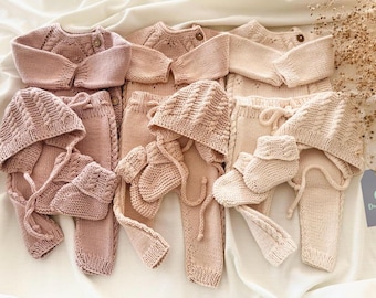 May include: Four knitted baby outfits in shades of pink and beige. Each outfit includes a hat, pants, and a sweater. The outfits are made with a soft, chunky yarn.