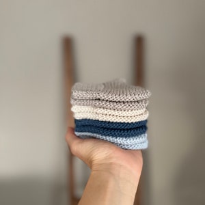 Może przedstawiać: Stoska dzianych ściereczek kuchennych w odcieniach beżowego i niebieskiego. Ściereczki są złożone i ułożone w dłoni.