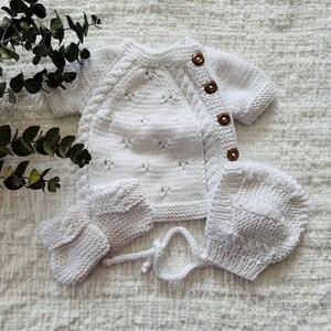 May include: A white knitted baby outfit set with a cardigan, hat, and booties. The cardigan has a button closure and a delicate stitch pattern. The set is made of soft, comfortable yarn.