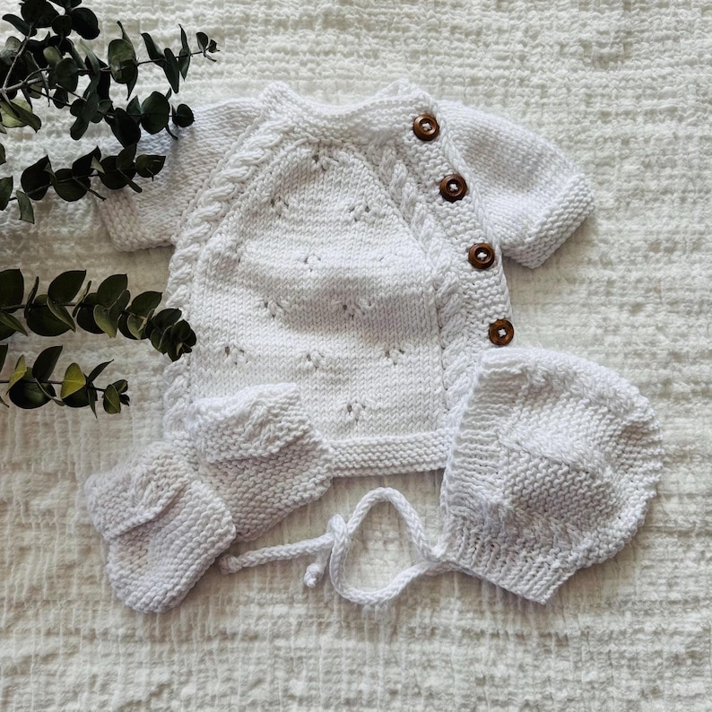 May include: A white knitted baby outfit set with a cardigan, hat, and booties. The cardigan has a button closure and a delicate stitch pattern. The set is made of soft, comfortable yarn.