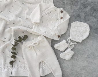 May include: A white knitted baby clothing set, including a blanket, cardigan, pants, booties, and bonnet. The set features a textured pattern and wooden buttons on the cardigan. The blanket measures approximately 30 x 40 inches.