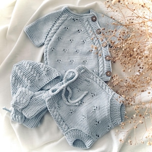 May include: A light blue knitted baby outfit set with a cardigan, pants, and a hat. The set features a delicate cable knit pattern and a button closure on the cardigan.