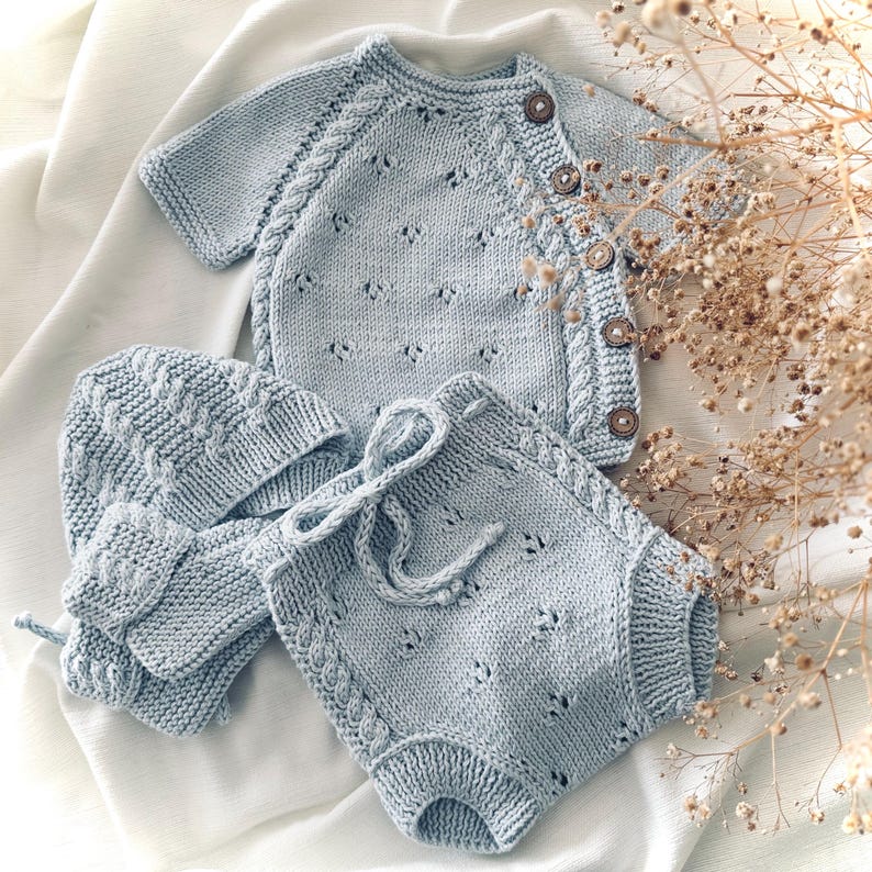 May include: A light blue knitted baby outfit set with a cardigan, pants, and a hat. The set features a delicate cable knit pattern and a button closure on the cardigan.