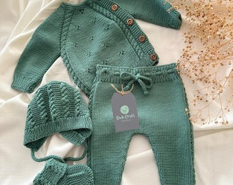 May include: A green knitted baby outfit set with a sweater, pants, hat, and booties. The set features a cable knit pattern and wooden buttons. The tag reads "Dodo Craft".