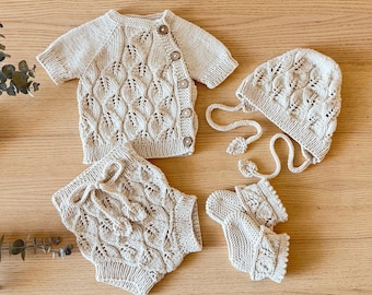 May include: A beige knitted baby clothing set, including a short-sleeved cardigan with wooden buttons, shorts with a drawstring, a bonnet with ties, and booties. The set features a leaf pattern.