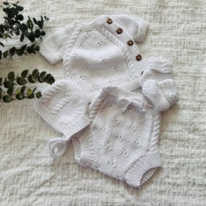 May include: A white knitted baby outfit set, including a sweater, pants, and a hat. The sweater has a button closure and a delicate lace pattern. The pants have a drawstring waist and a similar lace pattern. The hat is a simple beanie style.