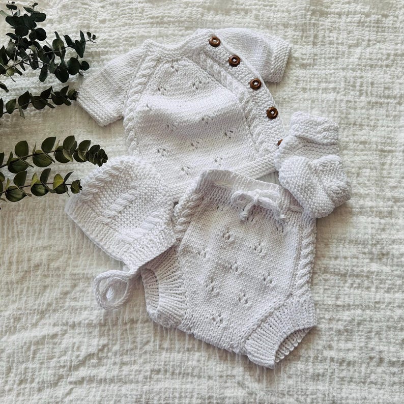 May include: A white knitted baby outfit set, including a sweater, pants, and a hat. The sweater has a button closure and a delicate lace pattern. The pants have a drawstring waist and a similar lace pattern. The hat is a simple beanie style.