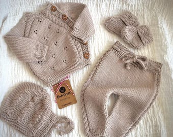 May include: A knitted baby outfit set in a light brown color. The set includes a cardigan, pants, a hat, and booties. The cardigan has a cable knit pattern and buttons down the front. The pants have a drawstring waist. The hat is a beanie style with a small brim. The booties are simple and have a fold-over cuff.