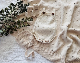 May include: A set of knitted baby clothes in a cream color, including a hat, a onesie, and booties. The clothes are laid out on a white fabric background with green leaves.