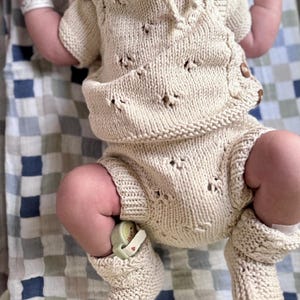 May include: A newborn baby wearing a knitted cream-colored outfit with a crocheted pattern. The outfit includes a short-sleeved top and shorts with matching booties.