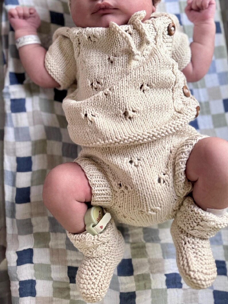 May include: A newborn baby wearing a knitted cream-colored outfit with a crocheted pattern. The outfit includes a short-sleeved top and shorts with matching booties.