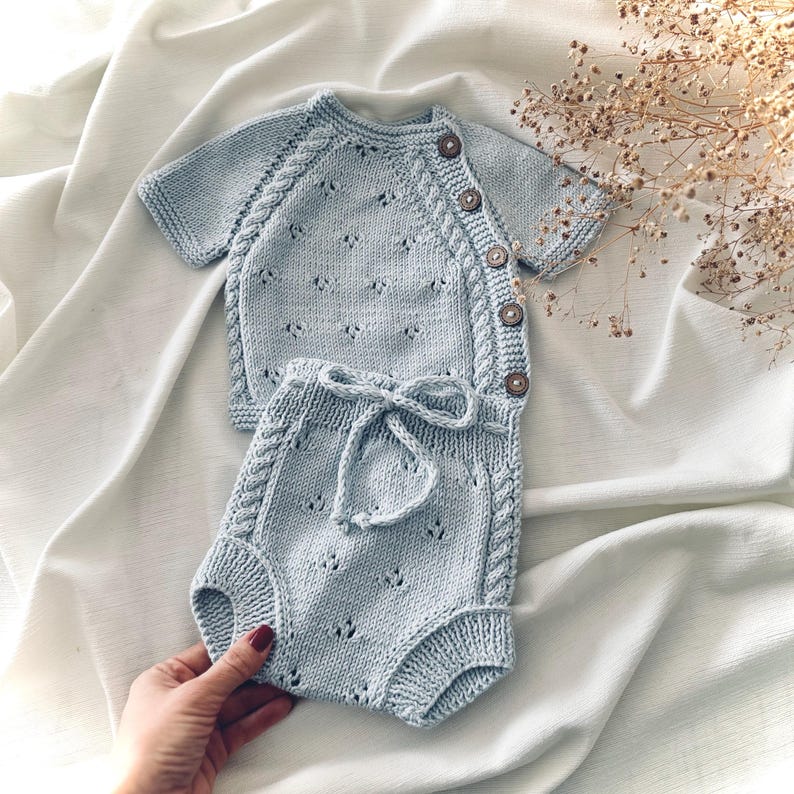 May include: A light blue knitted baby outfit with a short-sleeved top and matching pants. The top has a button closure and the pants have a tie at the waist.