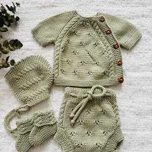 May include: A sage green knitted baby outfit set with a hat, booties, and shorts. The set features a delicate cable knit pattern and wooden buttons.