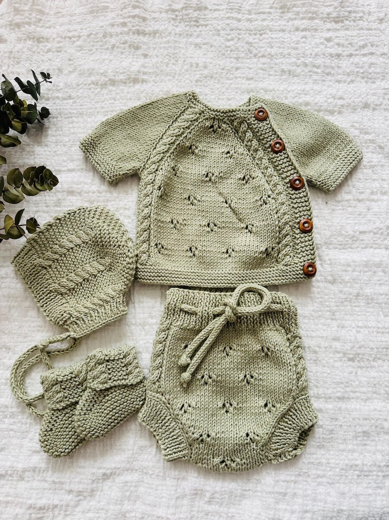 May include: A sage green knitted baby outfit set with a hat, booties, and shorts. The set features a delicate cable knit pattern and wooden buttons.