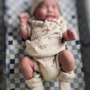 May include: A newborn baby wearing a cream-colored knitted outfit with a matching hat and booties. The baby is lying on a checkered blanket.