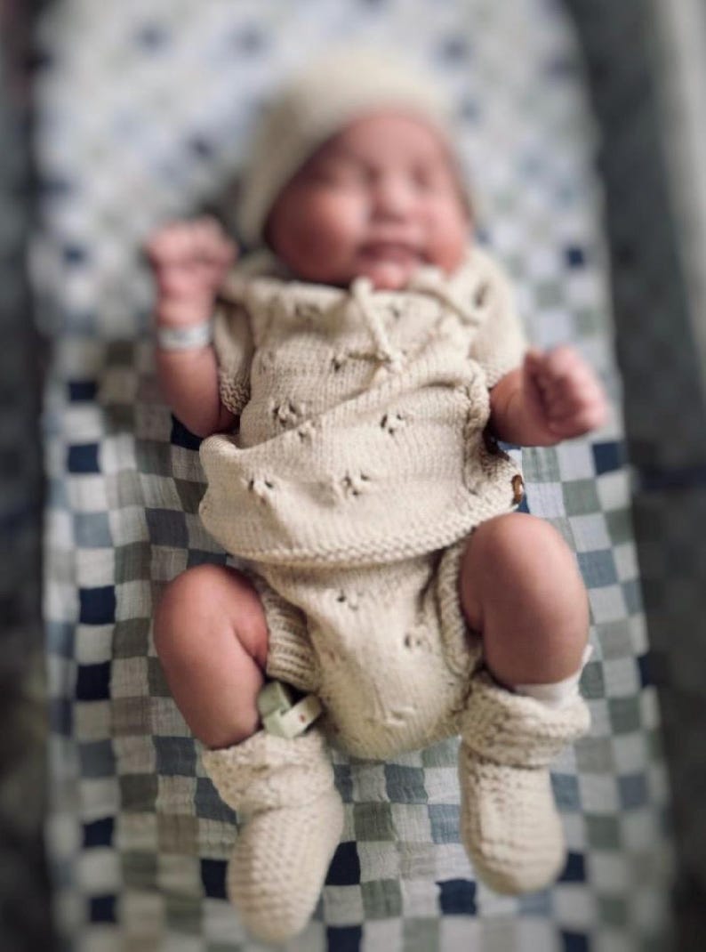 May include: A newborn baby wearing a cream-colored knitted outfit with a matching hat and booties. The baby is lying on a checkered blanket.