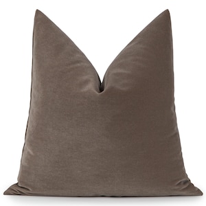 May include: A square, taupe-coloured cushion with a soft, velvety texture. The cushion has a classic shape with a pointed top, and is likely used for decorative purposes on a bed or sofa. The neutral colour makes it versatile for various interior design styles.