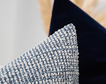 Peut inclure: Une housse de coussin texturée présentant un motif tissé de fils bleu clair et blanc cassé. Le tissu possède une surface granuleuse et tactile avec une construction en grille visible. Un coussin bleu marine uni est placé derrière.