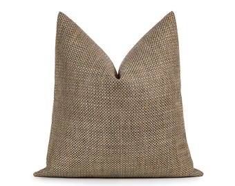 May include: A square throw pillow featuring a textured, woven fabric in a neutral brown and tan color palette. The material displays a consistent, small-scale grid pattern throughout, with a distinct crease at the top center of the pillow.