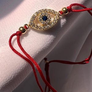 May include: A red string bracelet with a gold evil eye charm. The charm is decorated with clear rhinestones and a blue center stone.