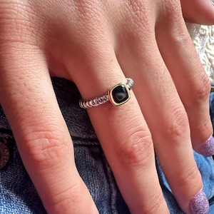 May include: A ring featuring a twisted silver band and a square-shaped black stone set in a gold-toned bezel. The design showcases a textured, cable-like metal band that wraps around the finger, highlighting the contrast between the dark center stone and the metallic frame.