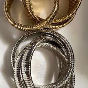 May include: Two sets of bracelets, one gold-toned and one silver-toned, are displayed. Each bracelet is composed of multiple, flexible, ribbed metal bands, creating a textured, snake-like appearance. The bracelets are arranged in overlapping loops.