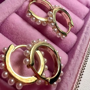 May include: Gold-tone hoop earrings adorned with small, round, white pearls. The earrings are displayed in a pink velvet jewelry box. The metal is stamped with "K925".