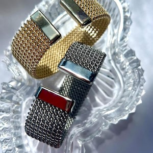 May include: Three cuff bracelets featuring a flexible mesh design. One bracelet is gold-toned with a polished gold-toned end cap, while two bracelets are dark grey with polished silver-toned end caps. The bracelets are arranged in a stack.