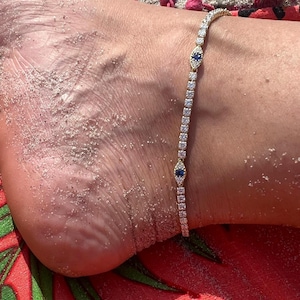 May include: A gold-toned anklet featuring a continuous row of clear, sparkling stones interspersed with two distinct eye-shaped charms. Each charm contains a central deep blue stone surrounded by a halo of small, clear, brilliant-cut stones.