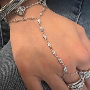 May include: A silver-toned hand chain featuring a delicate chain with marquise-cut clear stones connecting a wrist bracelet to a ring. The bracelet includes two clover-shaped charms. Additional stacked silver-toned rings and two thick wrist bangles.