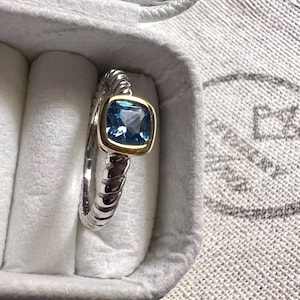 May include: A silver ring with a square, light blue gemstone set in a gold bezel. The ring band has a textured, ribbed design. The ring is displayed in a white jewelry box.