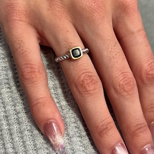 May include: A silver ring with a square black onyx stone set in a gold bezel. The band has a twisted rope design. The ring is on a hand with long, polished nails.