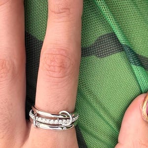 May include: A silver ring set with multiple bands, one with small clear stones. The ring has a knot design on the top band. The background is a green mesh fabric.